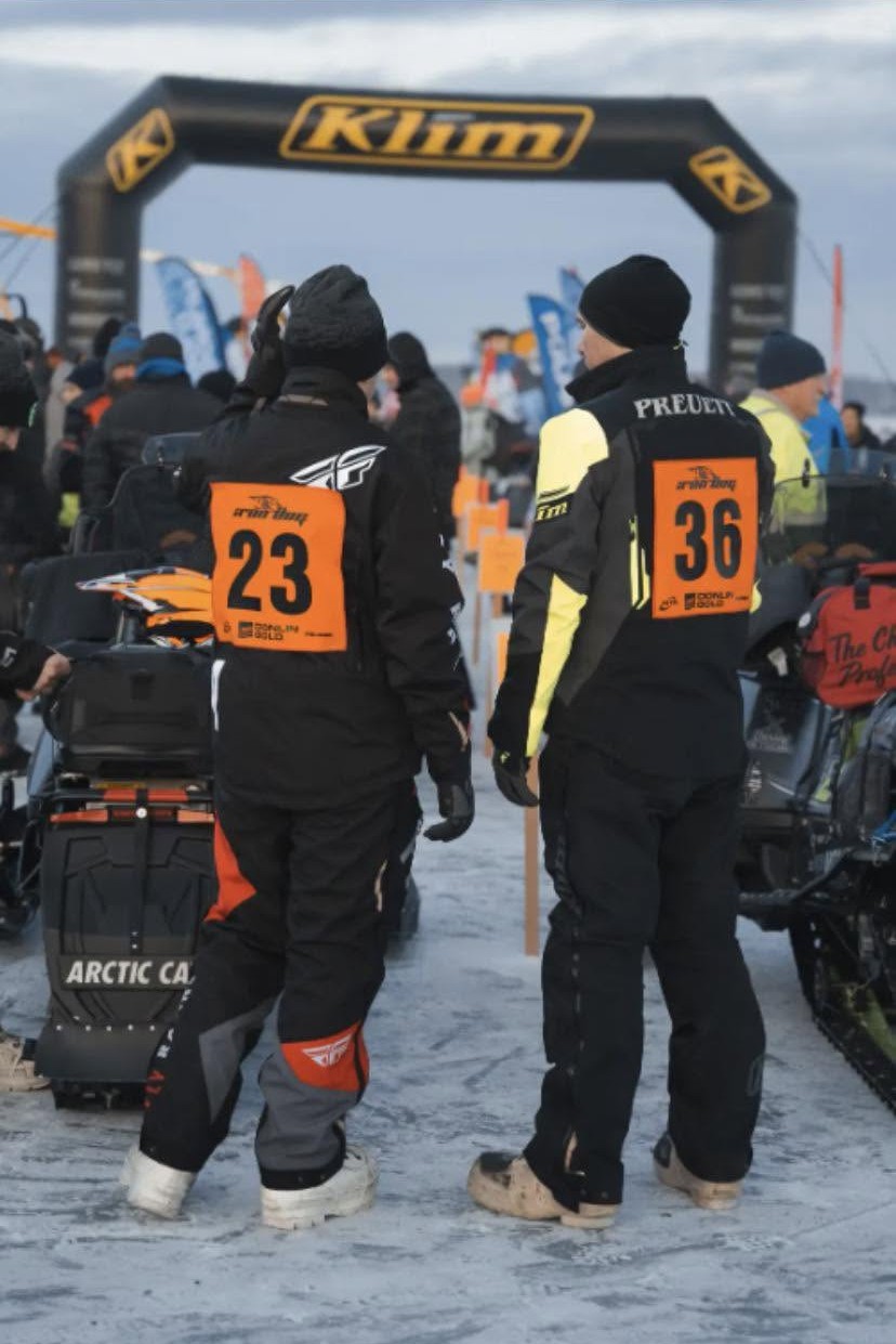 Iron Dog snowmobile racers wearing orange sublimated bibs 23 and 36 at the Klim start arch